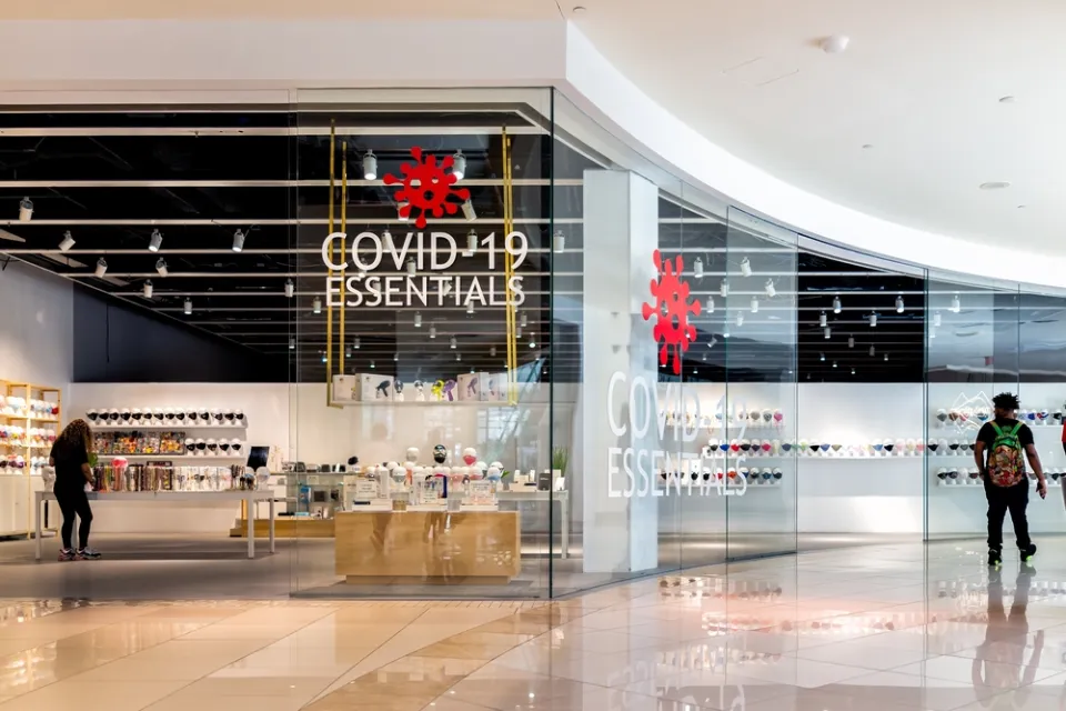 A man passes by a store in an empty mall. A sign in the window of the store reads "Covid-19 Essentials."